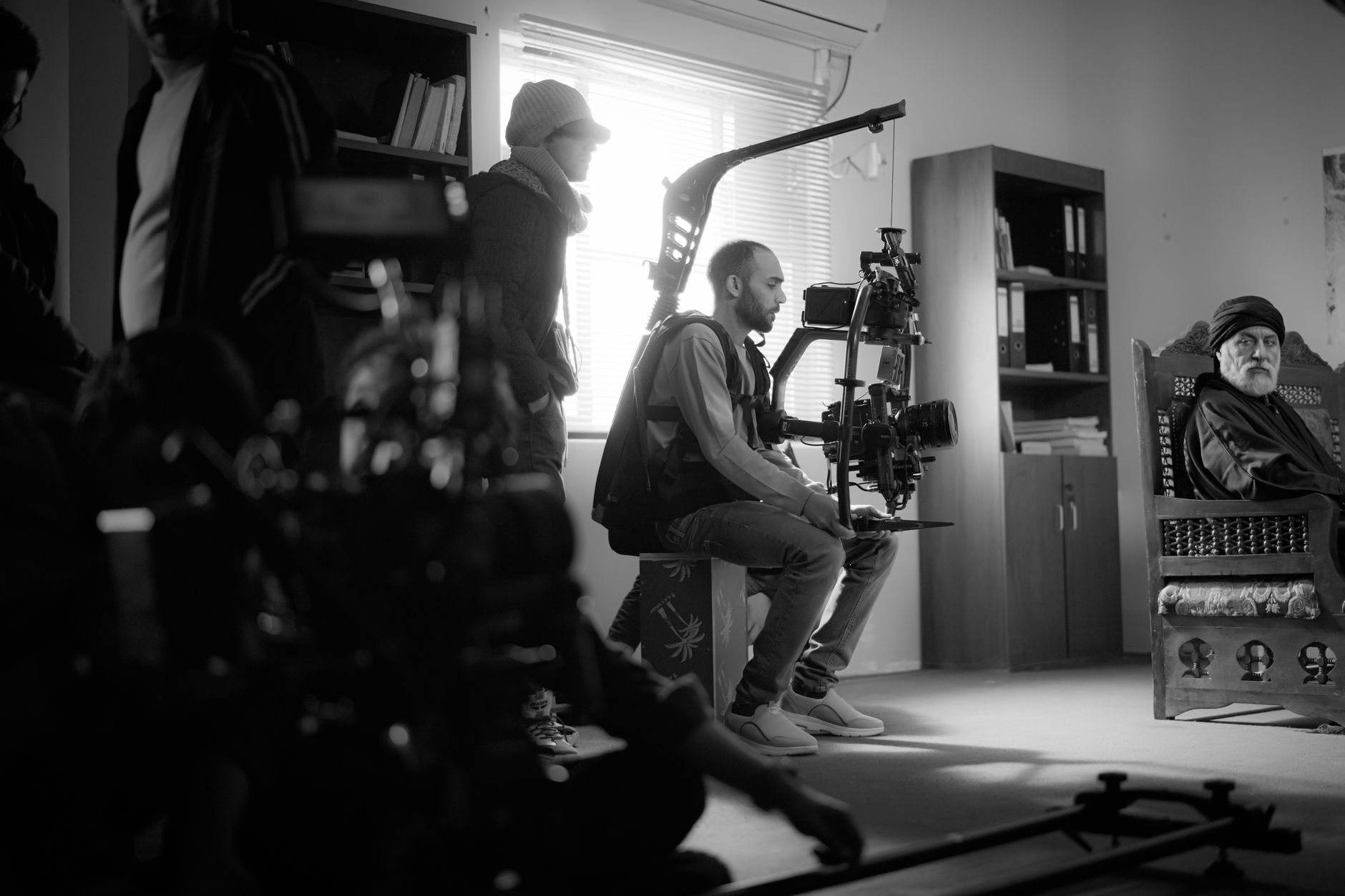 Équipe de tournage en noir et blanc : un opérateur assis tient une caméra sur stabilisateur, tandis qu’un homme barbu est assis dans un fauteuil à droite.