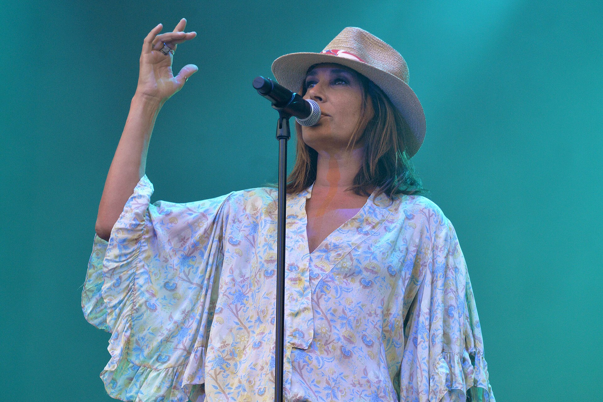 Zazie chante devant un micro sur pied, coiffée d’un chapeau de paille, sur un fond turquoise